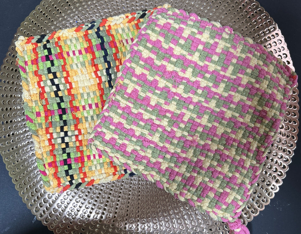 Two woven potholders, one multicolored and one with a checkered pattern, on a metal surface.